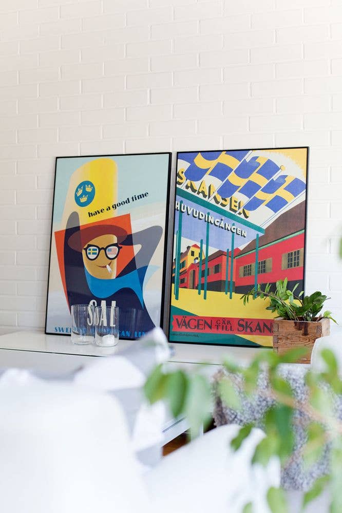 A framed vintage "Have a Good Time - Come to Sweden" poster is beautifully displayed on a shelf against a white brick wall, accompanied by a plant and glass decanter in the foreground. The high-quality matte paper enhances the elegance of the setup.