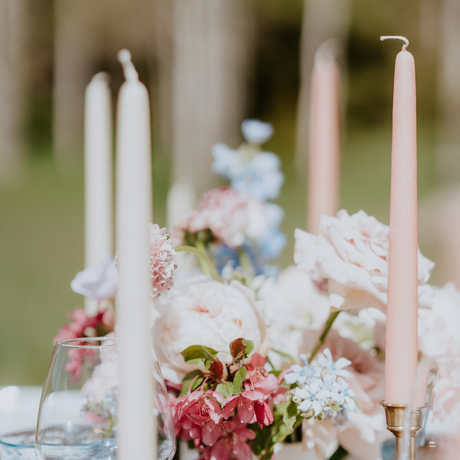 Eco-friendly, handmade beeswax taper candles in pink and white serve as dripless companions to a floral arrangement of pink, white, and blue flowers on a blurred outdoor table setting.