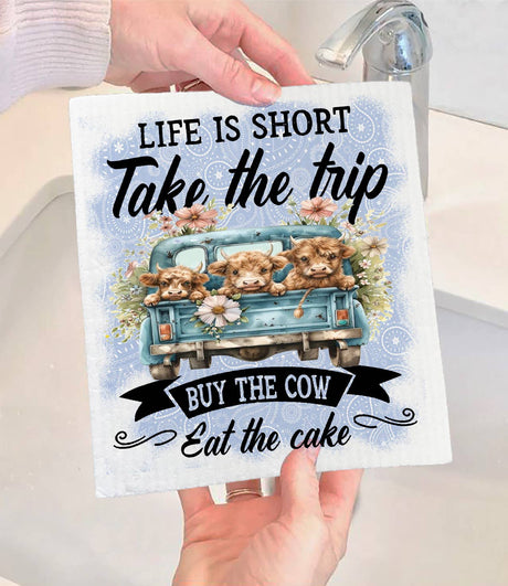 A person holds the Avery Lane Gifts Swedish Dishcloth, featuring three cows in a blue truck with flowers and the words: "Life is short. Take the trip. Buy the cow. Eat the cake." A sink and faucet are visible in the background.