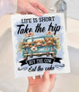 A person holds the Avery Lane Gifts Swedish Dishcloth, featuring three cows in a blue truck with flowers and the words: "Life is short. Take the trip. Buy the cow. Eat the cake." A sink and faucet are visible in the background.
