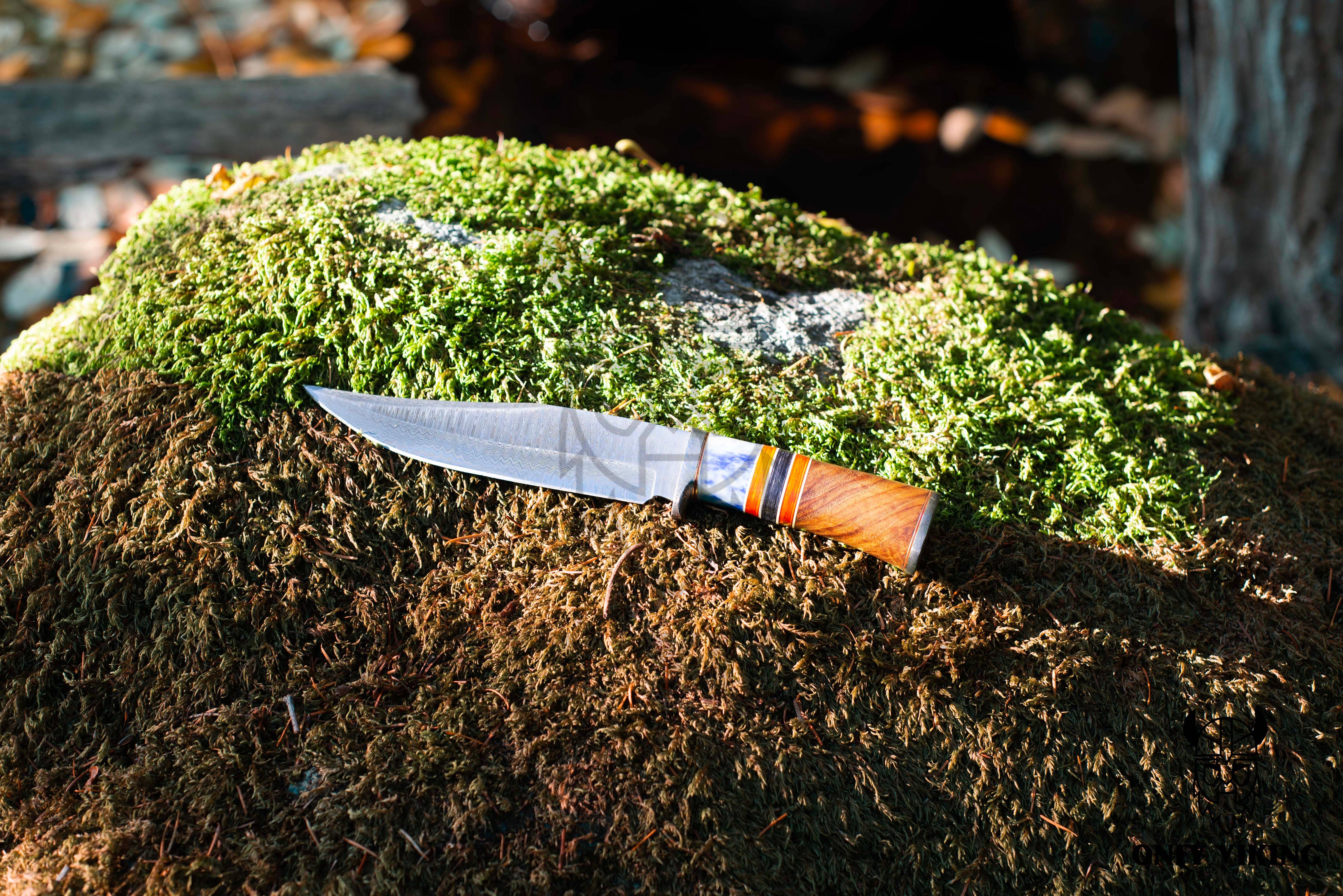 The Knife: Damascus Steel Hunting Knife, featuring a folding mechanism and a wooden handle with striped accents, rests on a moss-covered rock, showcasing its expertly crafted blade from Damascus steel.