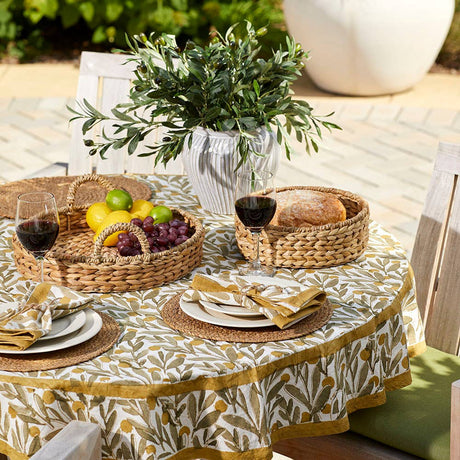 Outdoor table set for two with plates, wine glasses of red wine, a hand-braided fruit basket, bread served on a Natural Grass Braided Woven Placemat, and a potted plant centerpiece on a patterned tablecloth.