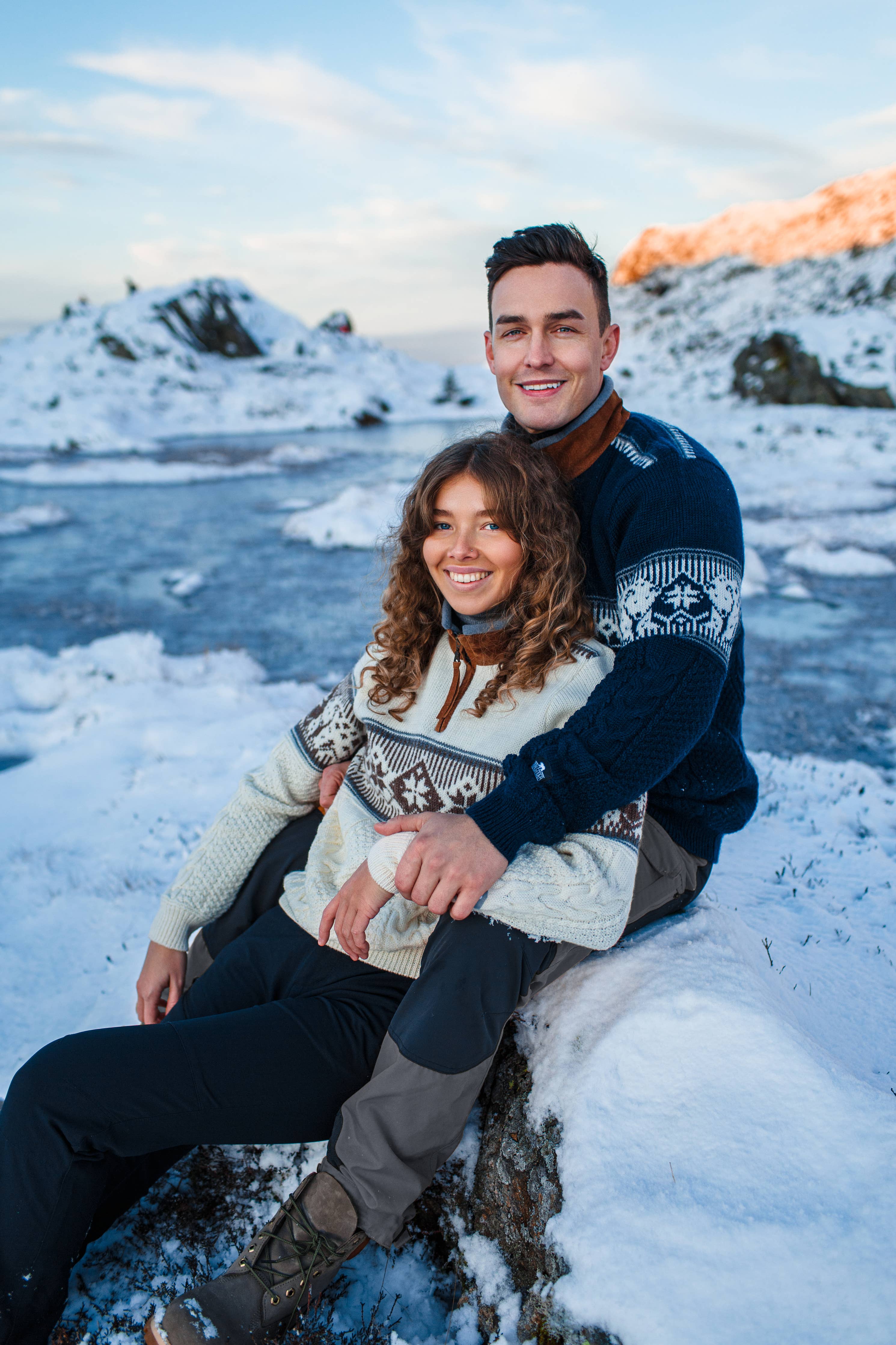 A couple in Spitzbergen Polar Sweater - Navy sits on a snowy landscape near a frozen lake, smiling at the camera with majestic mountains in the background.