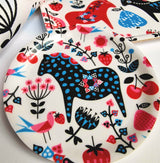 A decorative plate showcases a blue polka-dot Swedish Dala Horse, red cherries, pink flowers, and a pink bird on white—echoing the charm of Lotta Spjute’s Swedish Dala Horse Birchwood Coaster.