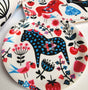 A decorative plate showcases a blue polka-dot Swedish Dala Horse, red cherries, pink flowers, and a pink bird on white—echoing the charm of Lotta Spjute’s Swedish Dala Horse Birchwood Coaster.
