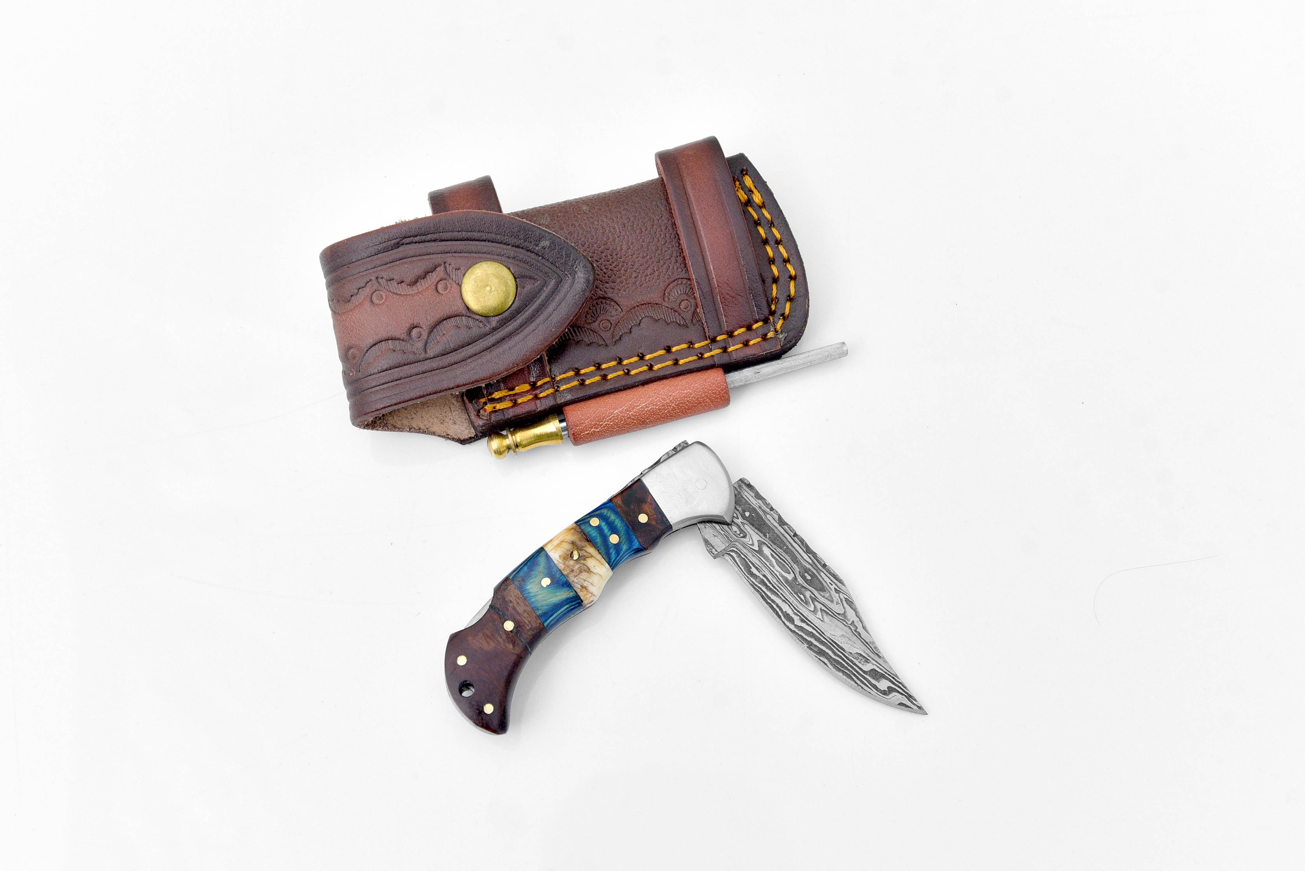 The Damascus Steel Pocket Knife, featuring a patterned blade and multicolored handle, rests beside a brown leather sheath with snap closure and decorative stitching.