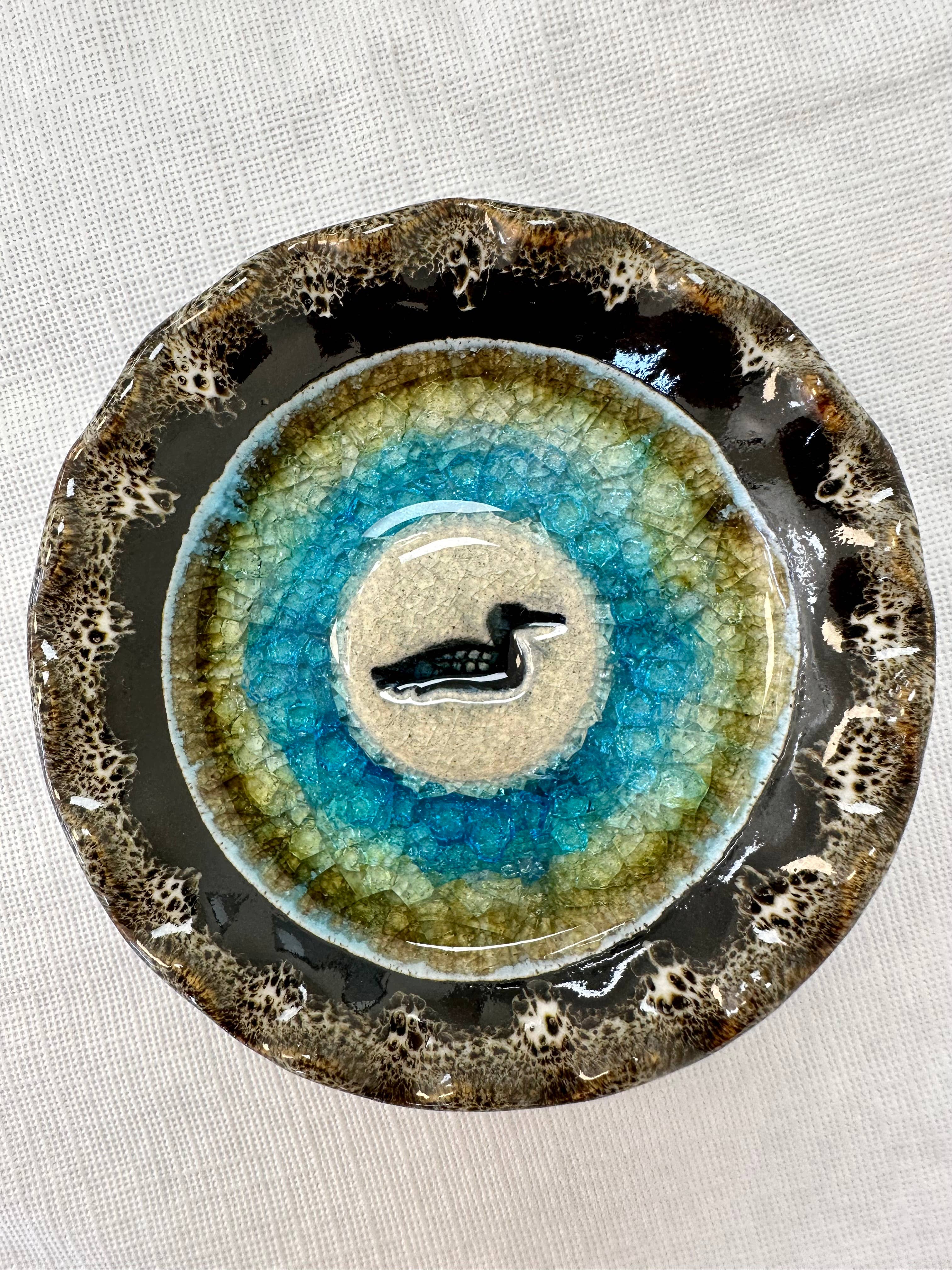 The Dish: Icon Series Loon Handmade Pottery is crafted from natural materials, featuring a crackled glaze in brown, green, and blue hues with a raised bird motif at the center.