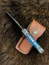 The Knife: Dyed Blue Bone 6.5 Damascus Steel, featuring a patterned blade and blue handle, rests on a brown leather sheath atop textured fur.