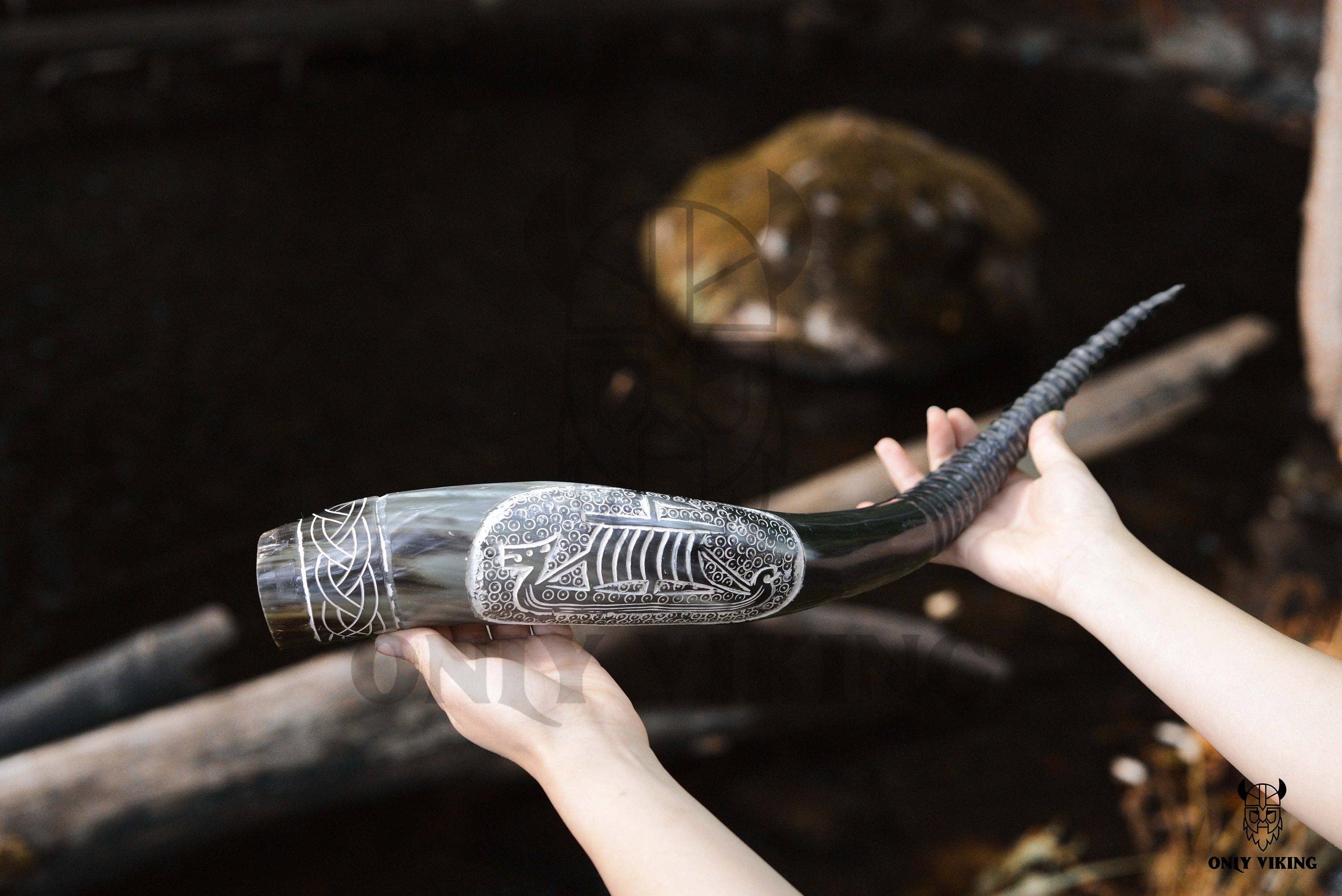 A person holds the Drinking Horn: Ravens Viking Ship Handcrafted Drinking Horn (14-18 inches) with an ornately carved ship design and metal stand, set against a dark, blurred background.