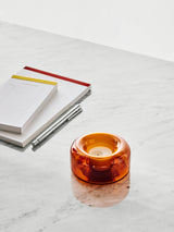 Amber blown glass votive candle holder on a white marble surface, positioned next to a stack of notepads and a silver pen.