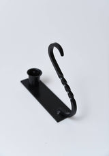 Iron taper holder with a rectangular base, circular candle cup, and a twisted S-shaped wall-mounting hook.