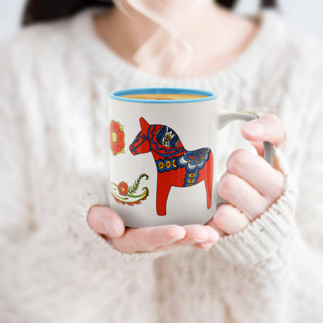 A person in a white sweater holds a steaming Coffee Mug: Red & Blue Dala Horses Ceramic Coffee Mug – Blue Interior (12 oz), featuring colorful Scandinavian folk art and red Dala horses design.