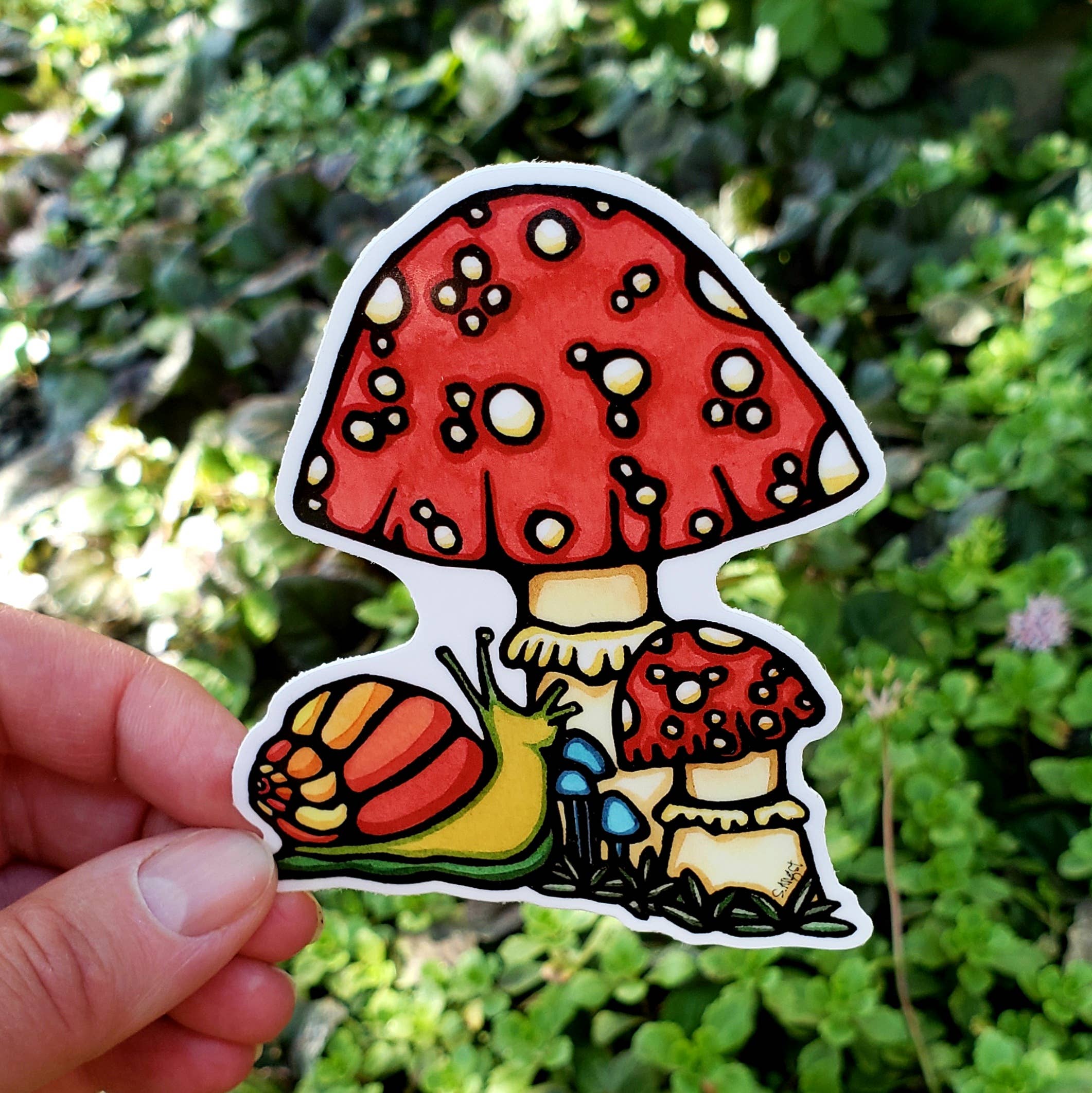 A hand holds the vibrant Mushroom Vinyl Sticker, featuring a yellow and orange snail near three red-spotted mushrooms amid greenery—a perfect find for any nature lover.