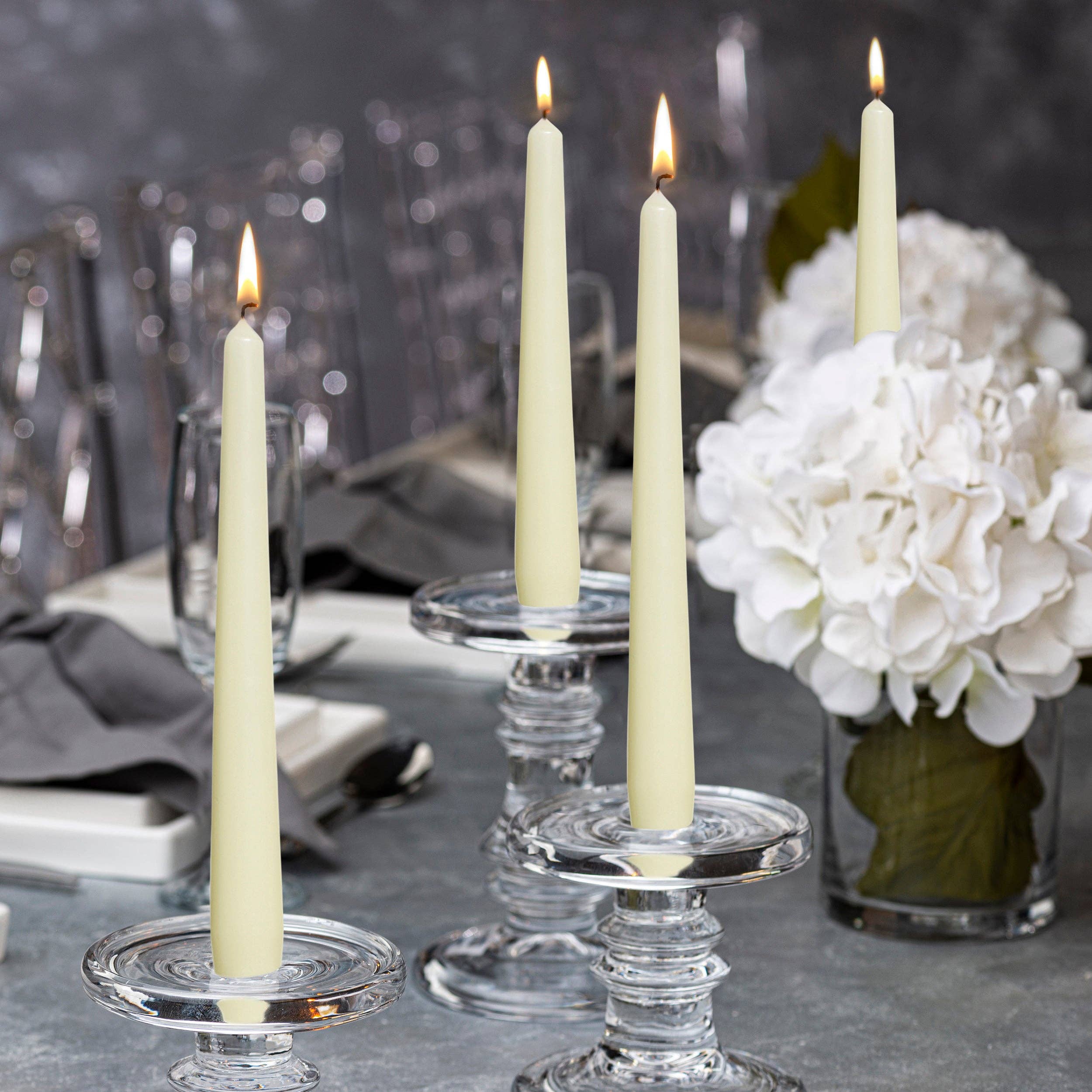 Four 10" tall white taper candles, designed for a flawless burn using high-quality paraffin wax, stand proudly in clear glass holders on a dining table. Accompanied by gray napkins and a beautiful white flower arrangement, these unscented candles create an inviting atmosphere without drips.