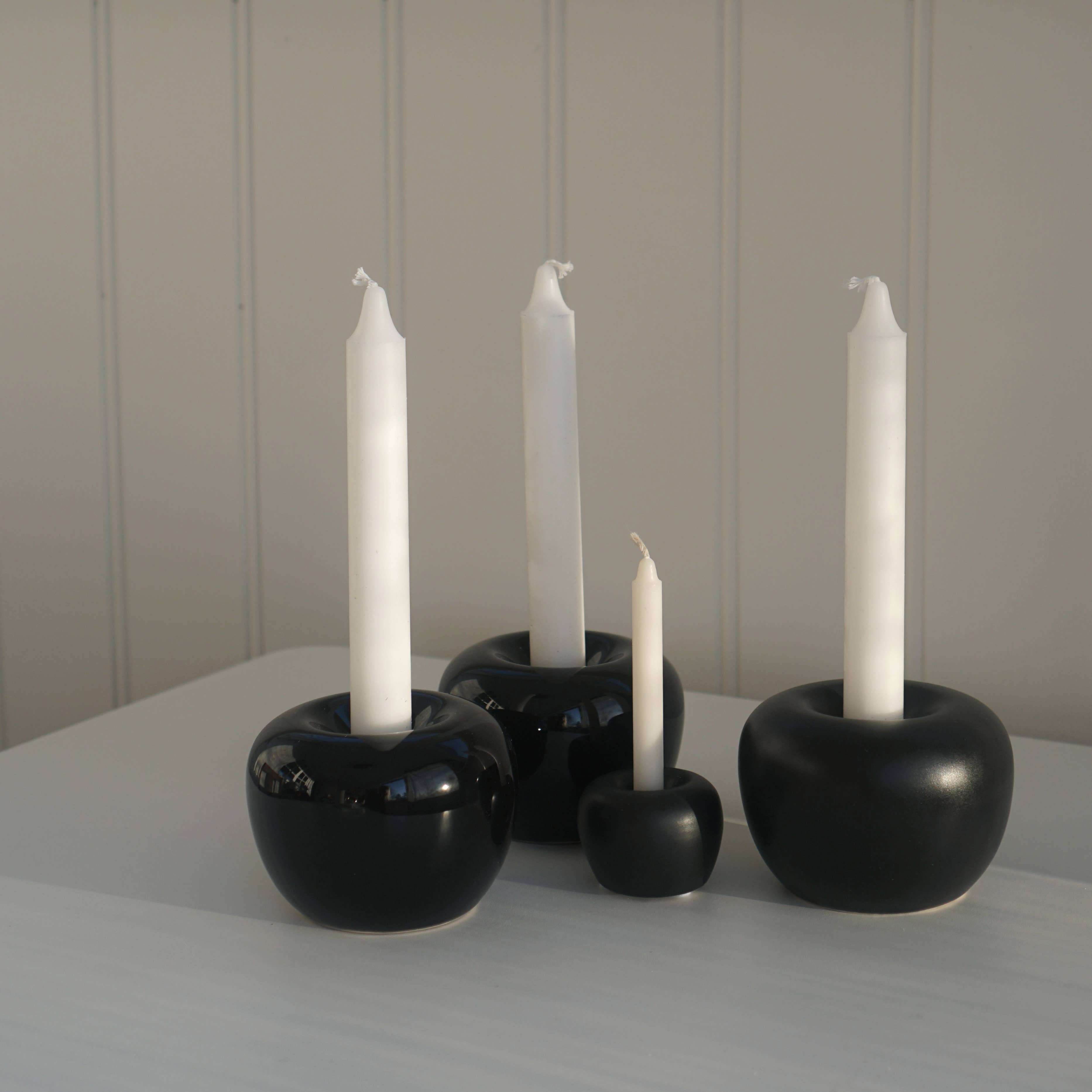 Four unlit white candles in stylish mini apple-shaped black candle holders on a stoneware surface against a light-paneled background.