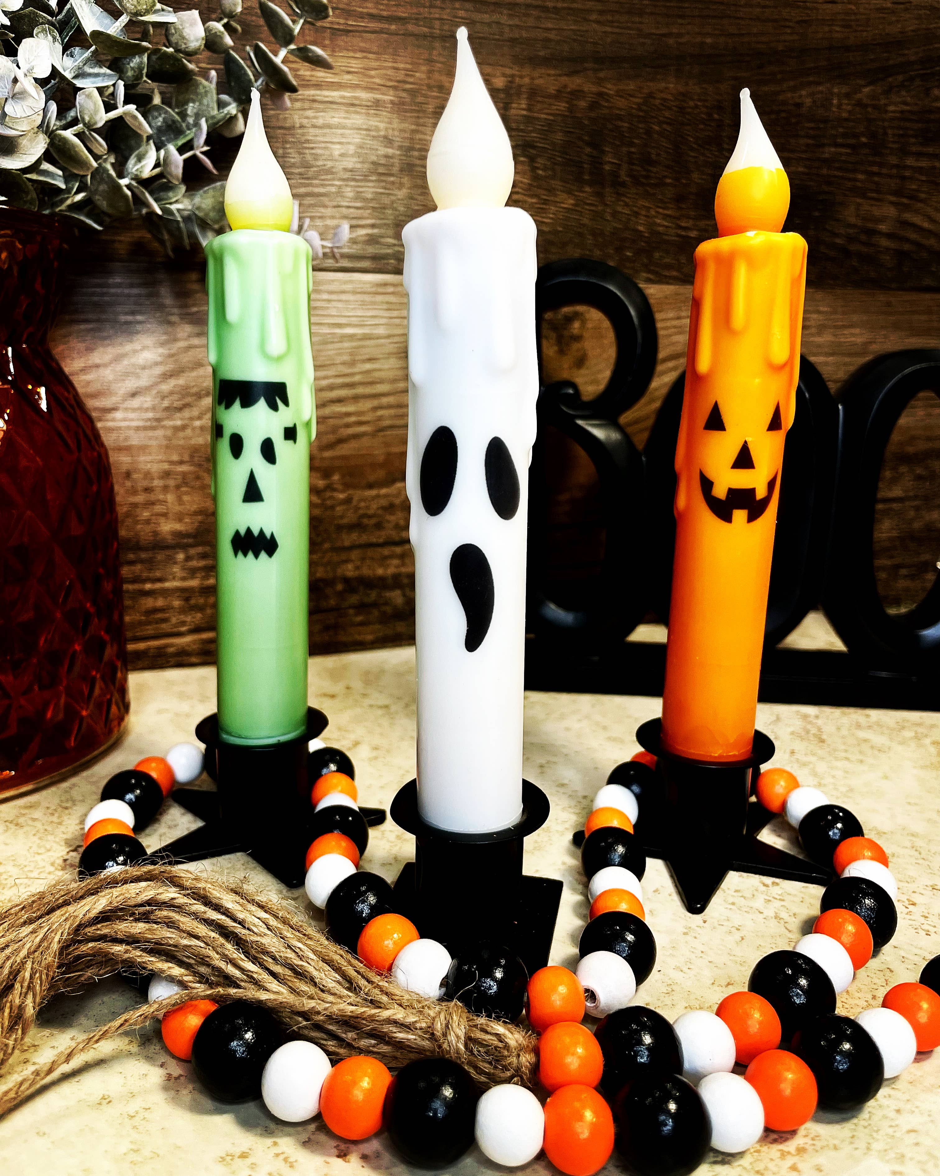 A trio of Halloween candles includes a Jack-O-Lantern, monster, and ghost design, each with colorful beads and twine in the foreground. These battery-operated taper candles capture the spooky essence while providing an authentic glow to complement your eerie decor.