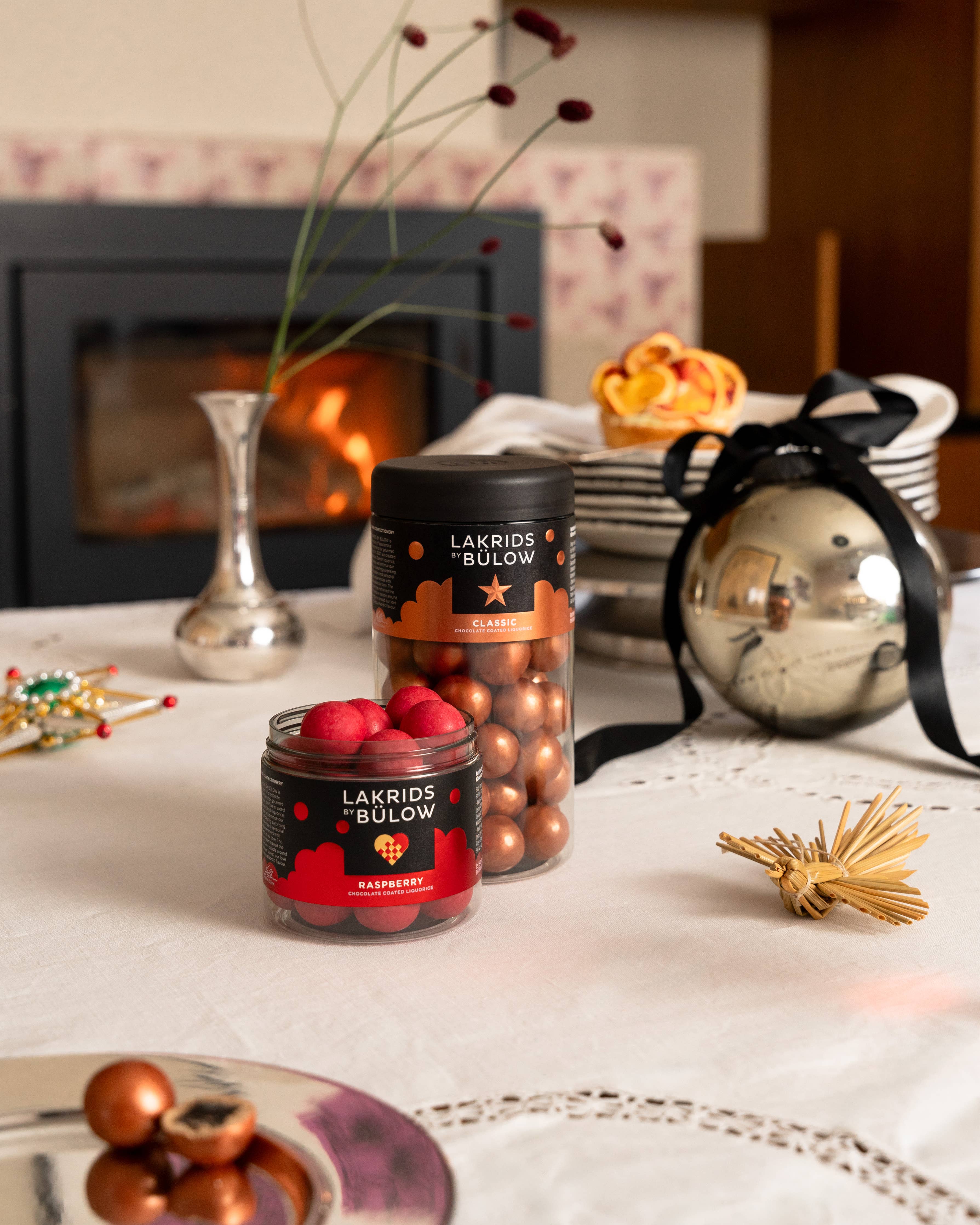 A jar of Lakrids by Bülow CLASSIC CARAMEL Chocolate Coated Licorice, Regular Limited Edition, sits on a decorated table near a fireplace and festive ornaments.