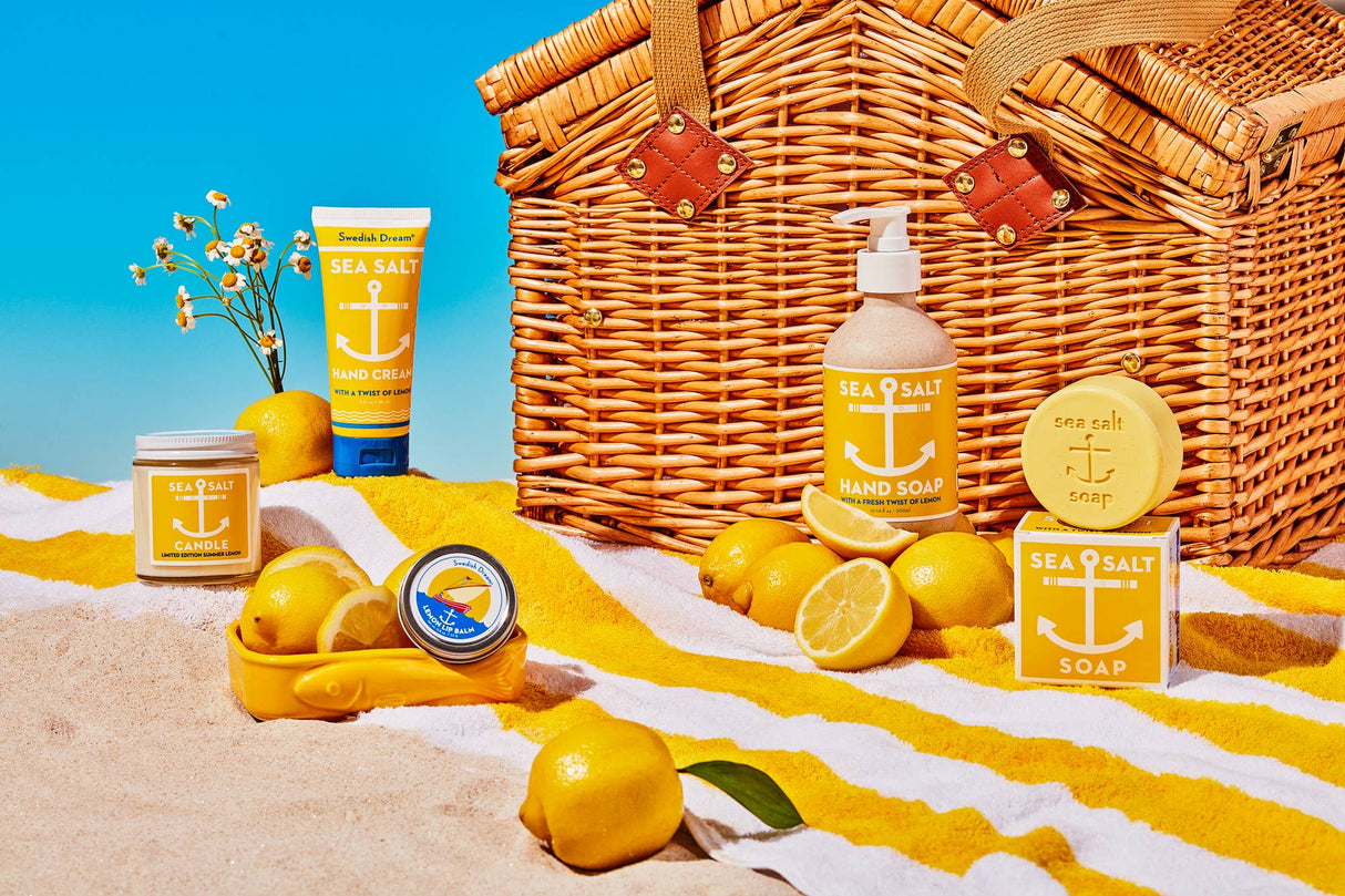 A wicker picnic basket and yellow-striped towel on a sandy beach display Swedish Dream Sea Salt Lemon Soap (4 oz), hand cream, a candle, and lemons beneath a clear blue sky.