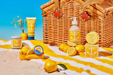 A wicker picnic basket and yellow-striped towel on a sandy beach display Swedish Dream Sea Salt Lemon Soap (4 oz), hand cream, a candle, and lemons beneath a clear blue sky.