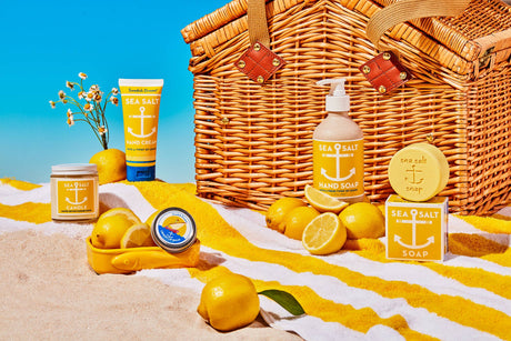 A wicker picnic basket and Swedish Dream Sea Salt Lemon Soap are displayed on a striped towel with lemons and daisies, set on sandy beach under a blue sky.