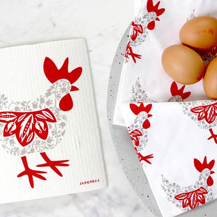 A Swedish dish cloth, designed in Scandinavia and featuring a hen in grey and red, is displayed next to three brown eggs on a plate.