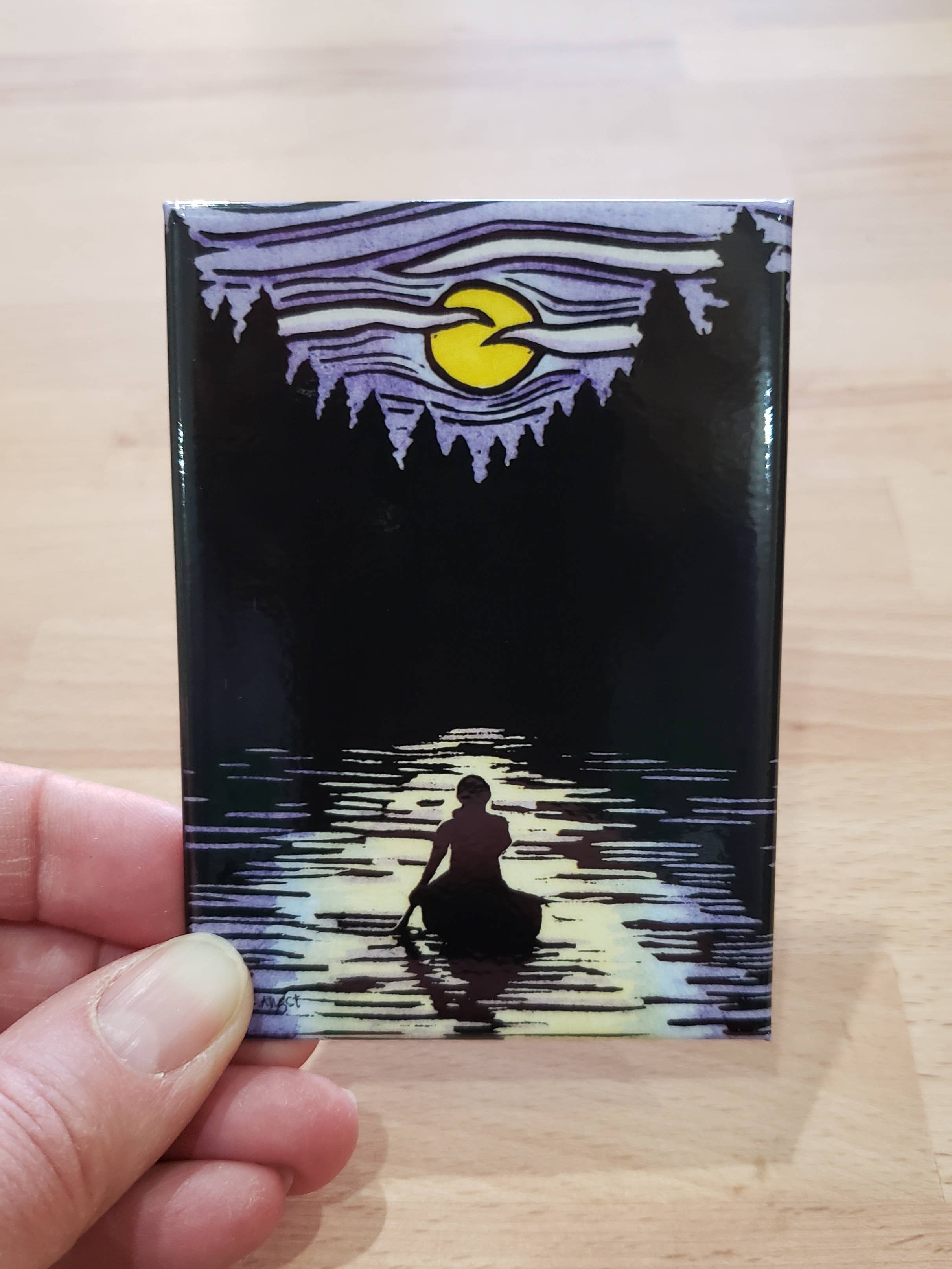 A hand holds the Solitude Canoe Magnet, a small artwork of a silhouette in a canoe on a lake, beneath wavy striped sky and crescent moon, framed by the forest, evoking an enchanting nighttime scene.