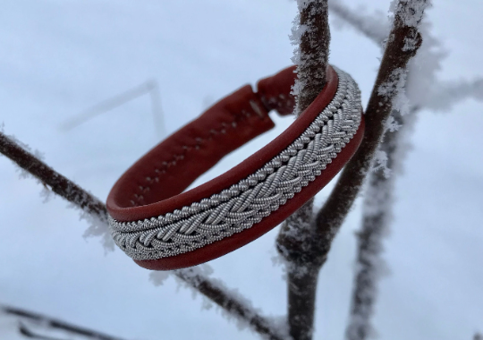 The Lapland Sami Tennis Bracelet #2 in cognac (19-20 cm), handmade in Sweden with braided reindeer leather and silver detailing, hangs on a frost-covered branch against a snowy backdrop.