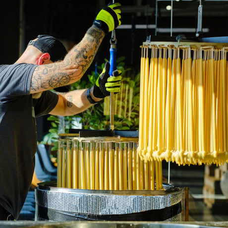 In a factory, a tattooed worker with gloves carefully crafts Beeswax Taper Candles, ensuring each eco-friendly 10 candle remains dripless for a cleaner and sustainable burn.