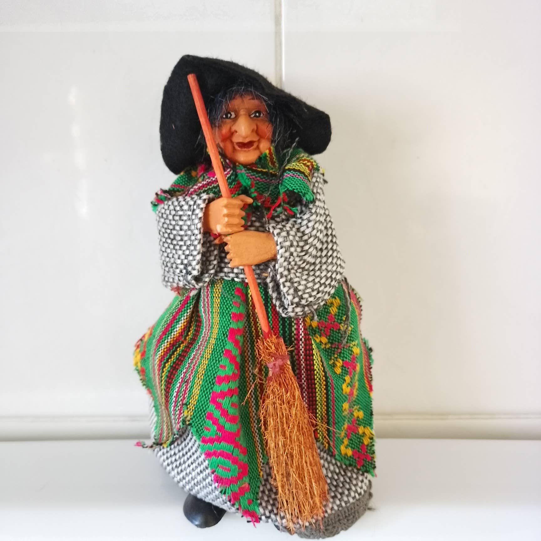 The Yasmina Green Kitchen Witch Figurine, 7" tall, features an elderly woman in a headscarf and patterned clothes holding a broom, standing against a plain white background.