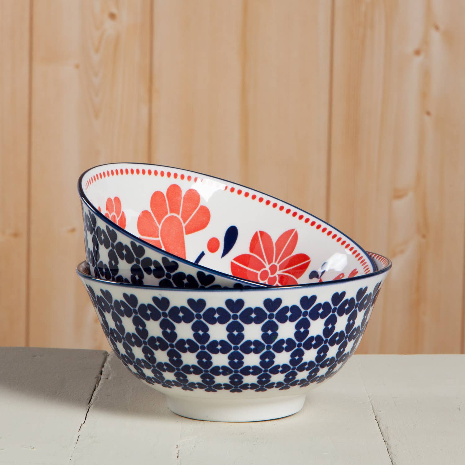 Two stacked Navy Bird Stamped Bowls, featuring red and blue floral patterns, rest on a wooden surface, exuding a subtle cottage chic charm.