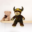 A small Viking Boy Plush Teddy Bear in a horned helmet and overalls sits on a shelf, making an adorable nursery companion or unique Oktoberfest gift.