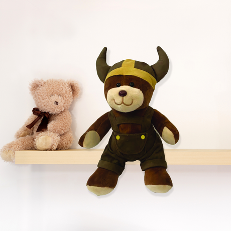 A small Viking Boy Plush Teddy Bear in a horned helmet and overalls sits on a shelf, making an adorable nursery companion or unique Oktoberfest gift.