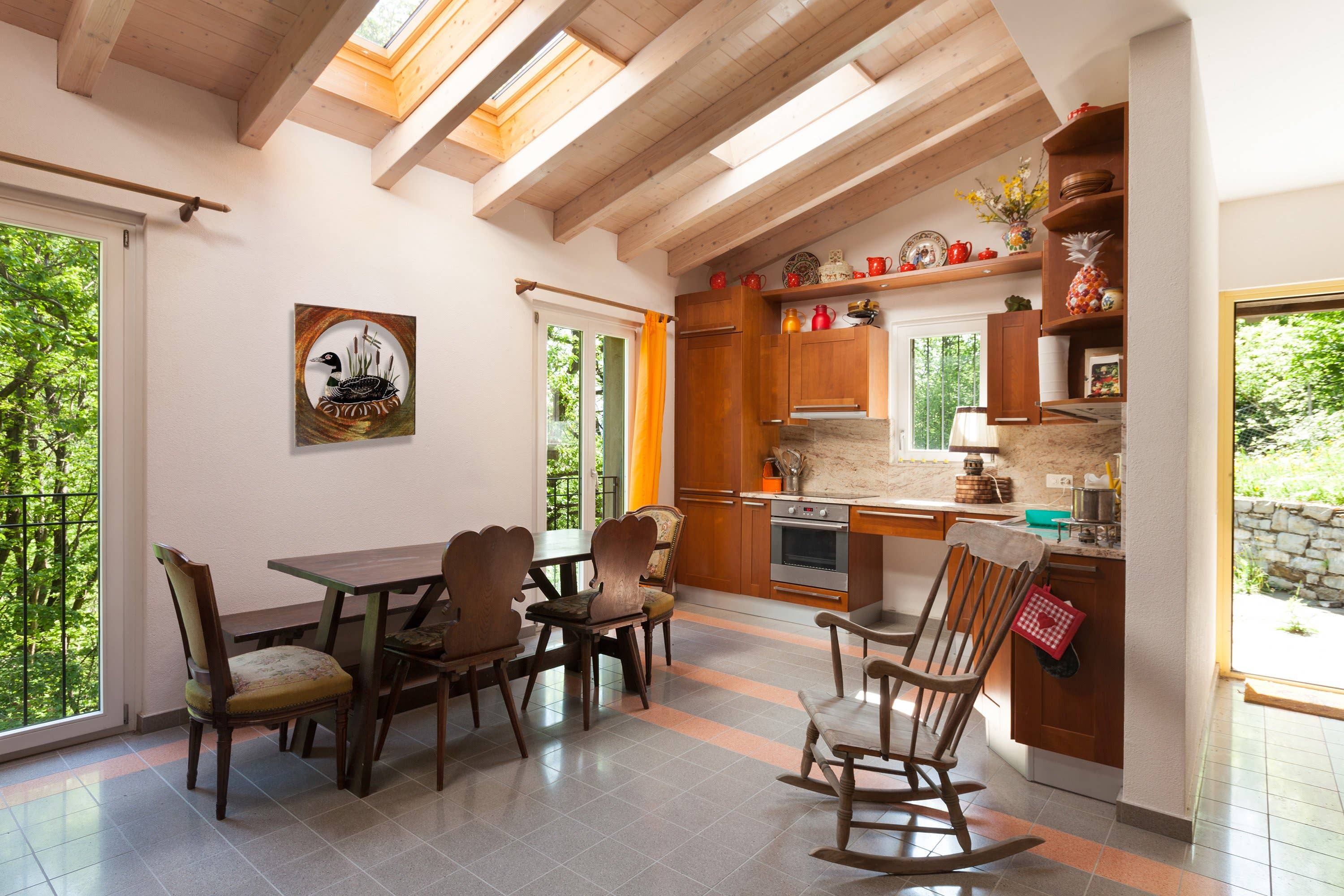 A cozy kitchen with wooden cabinets features a dining table and chairs, where sunlight streams through skylights and a door opens to a garden. The Loon Scene Metal Wall Art beautifully complements the decorative items on the shelves, adding charm to this inviting space.