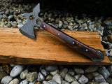 Patterned Damascus steel axe with a polished wooden handle resting on a wooden plank, surrounded by assorted stones.