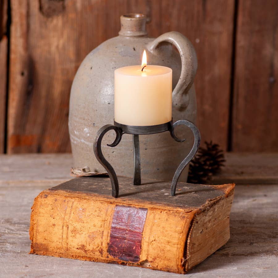 A lit candle on the Raised Wrought Iron Ball Candle Holder sits atop a worn book; a ceramic jug and pinecone are set against a rustic wooden background.