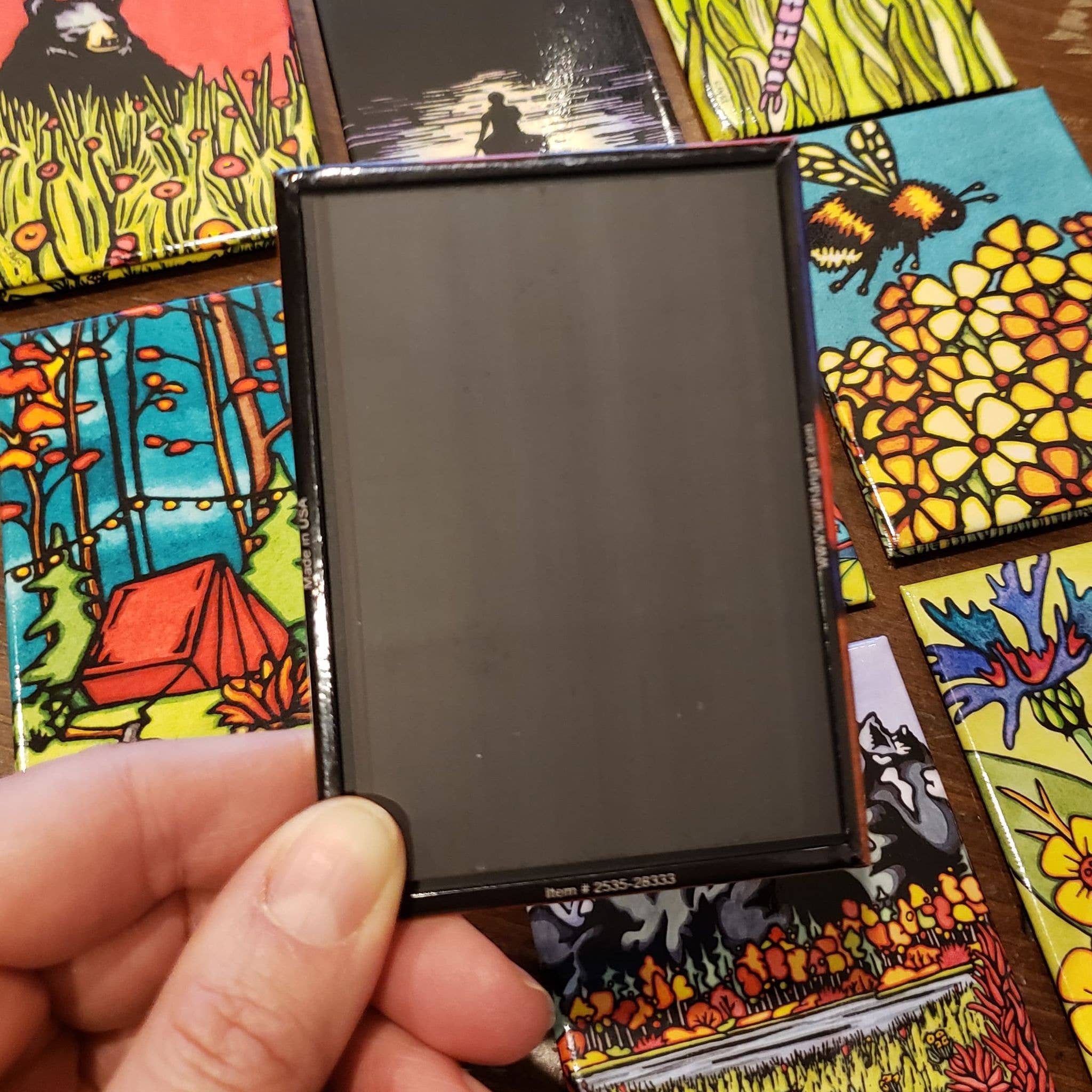 A hand holds a small black frame surrounded by colorful nature-themed cards with flowers, bees, and a tent—all highlighted with the Forest Moose Magnet.