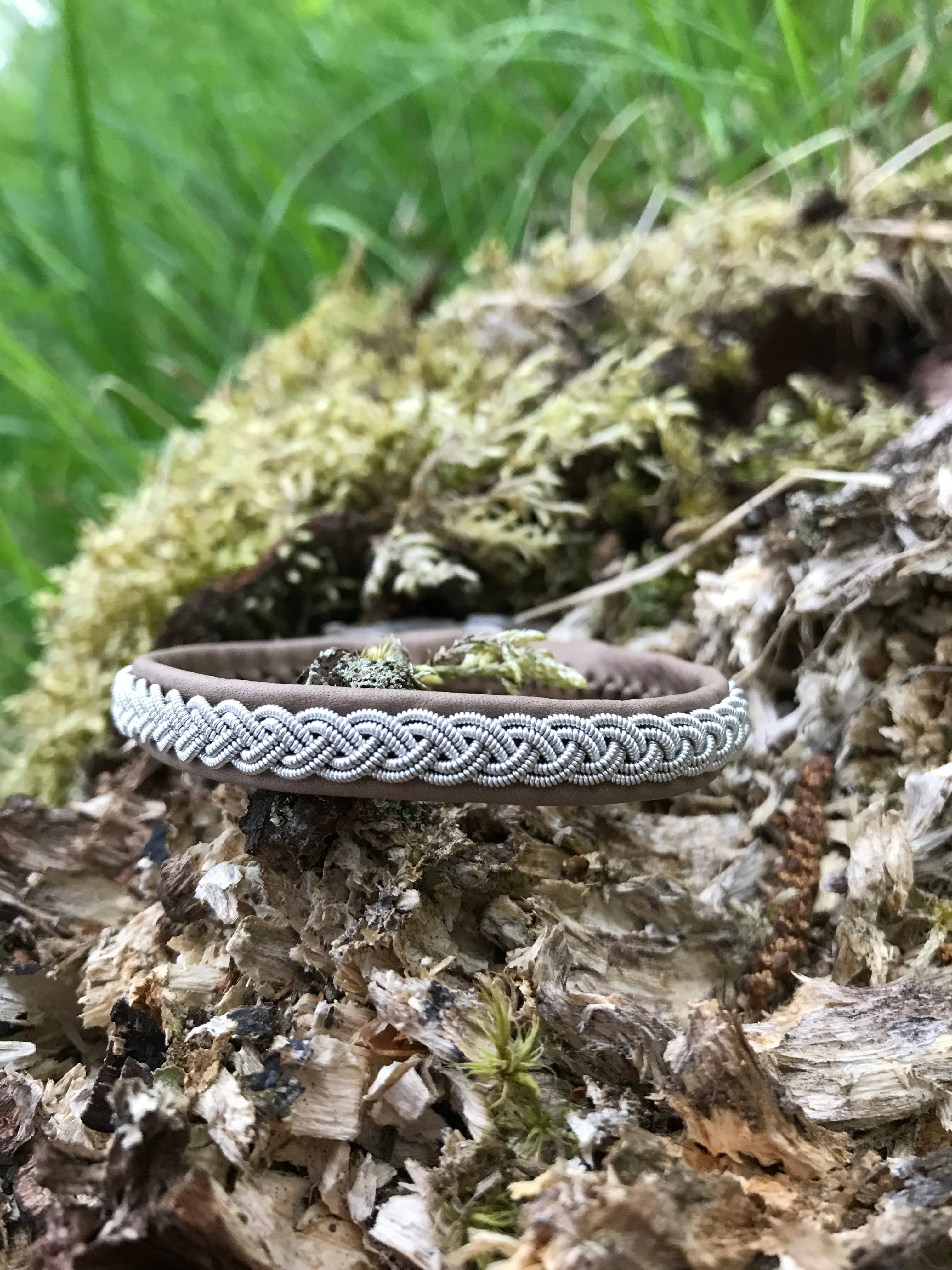 The Lapland Sami Tin Bracelet Single Braid #1 in blue gray rests on moss-covered wood with green grass behind, highlighting the distinctive charm of handmade Lapland jewelry.