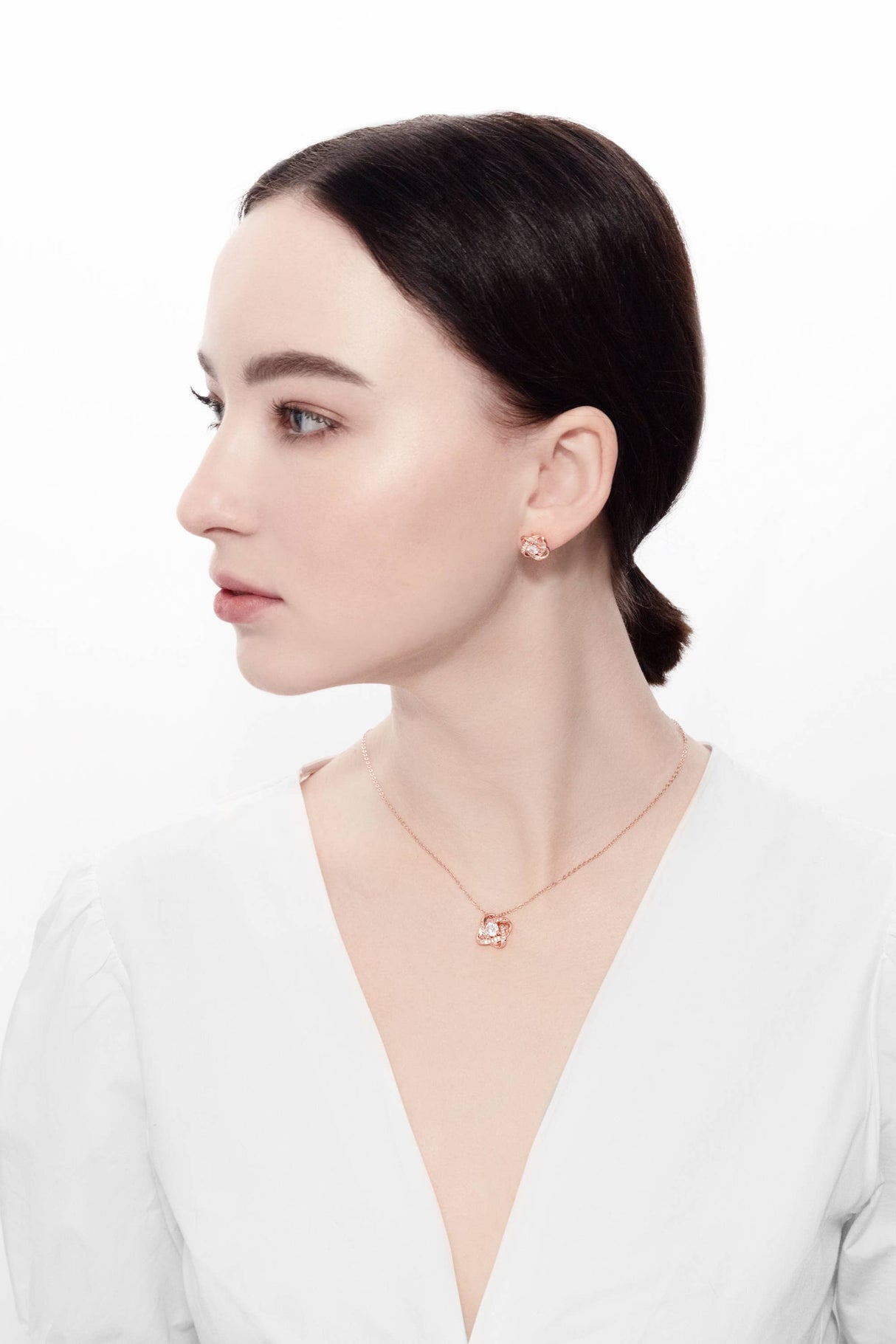 A woman with dark hair in a low bun wears a white top, rose gold earrings, and the Ice Dance Rose Gold/Sterling Silver Necklace with Brilliant Cut Cubic Zirconia, looking to the side against a plain white background.