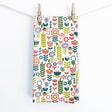 The Juniper Blue "Tuli" Scandinavian Floral Folk-Art Tea Towel (19x27 in) with hanging tab, featuring bright stylized flowers and leaves, hangs from a white clothesline with two wooden clothespins against a plain background.