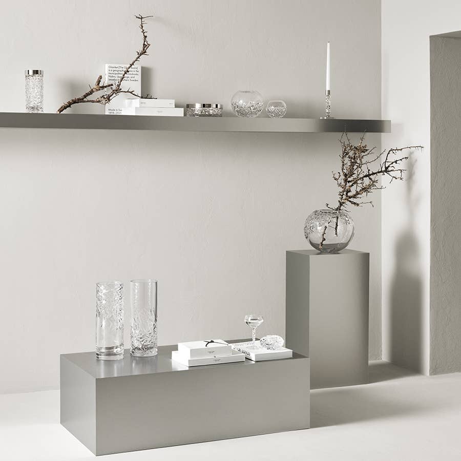 Gray shelves with a Carat Votive candle holder, clear glass vases, bare branches, books, and decorative items arranged neatly against a light-colored wall.