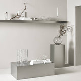 Gray shelves with a Carat Votive candle holder, clear glass vases, bare branches, books, and decorative items arranged neatly against a light-colored wall.