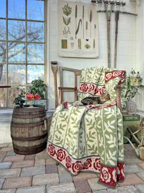 A cat lounges on a Kerstin Landstrom Rose Garden blanket made of 100% Norwegian lambs wool, draped over a wooden chair. Nearby, a barrel table with apples sits beside a rake and plant poster. A large window reveals the tree outside, casting gentle light on this serene scene.