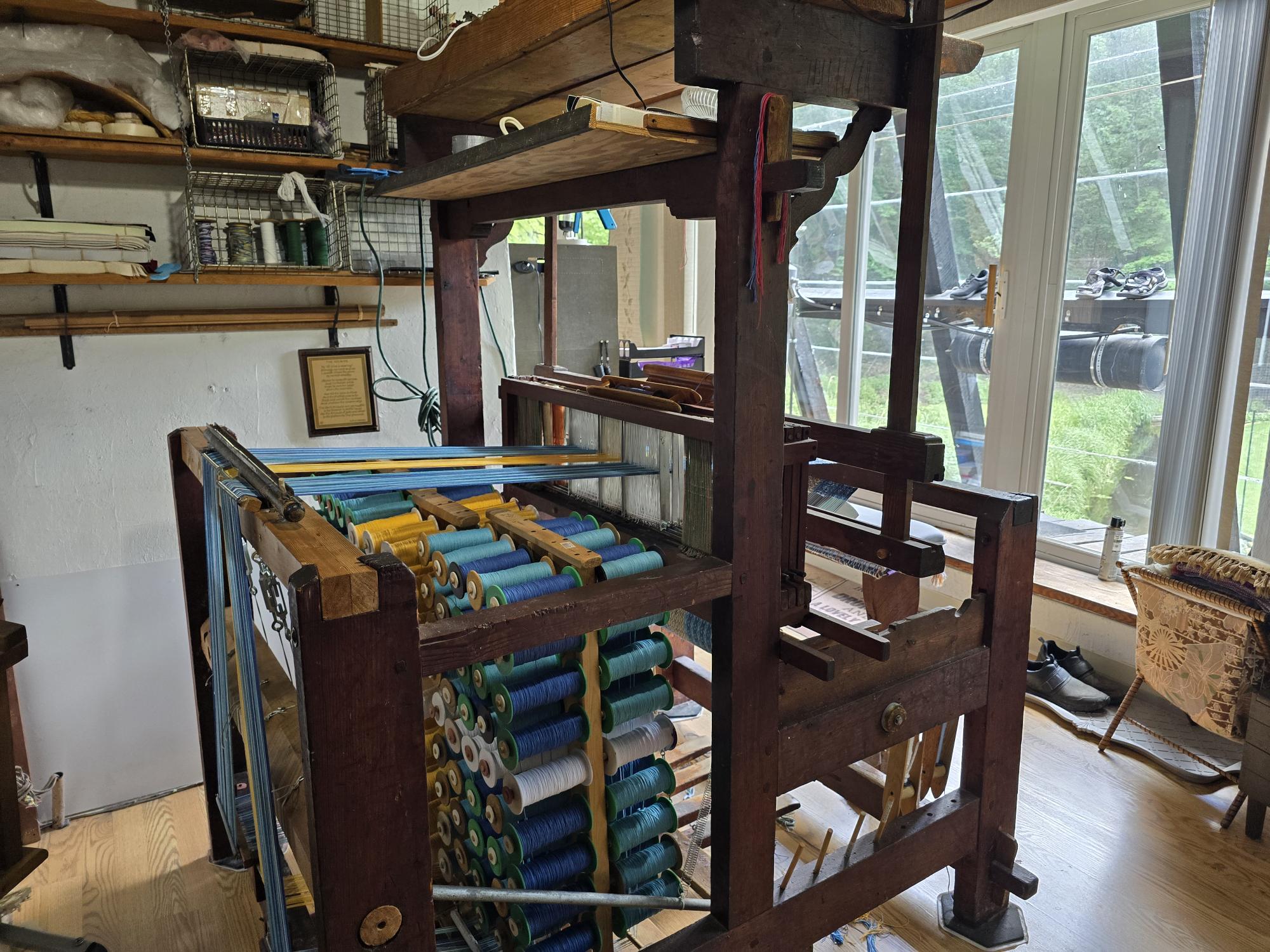 A vintage wooden loom with colorful threads, featuring a Swedish Flag Runner in 100% cotton and measuring between 46" and 49" in length, is set up near large windows. Shelves filled with supplies are visible in the background, showcasing the artistry of hand-woven crafts.