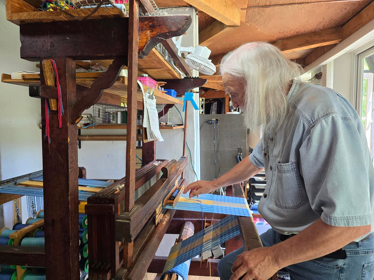 An older artisan hand-weaves the Center Piece: Swedish Flag 11\x12\ Plus Fringe on Long Ends from 100% cotton blue and white threads, reminiscent of the Swedish flag, using a wooden loom in a bright workshop.
