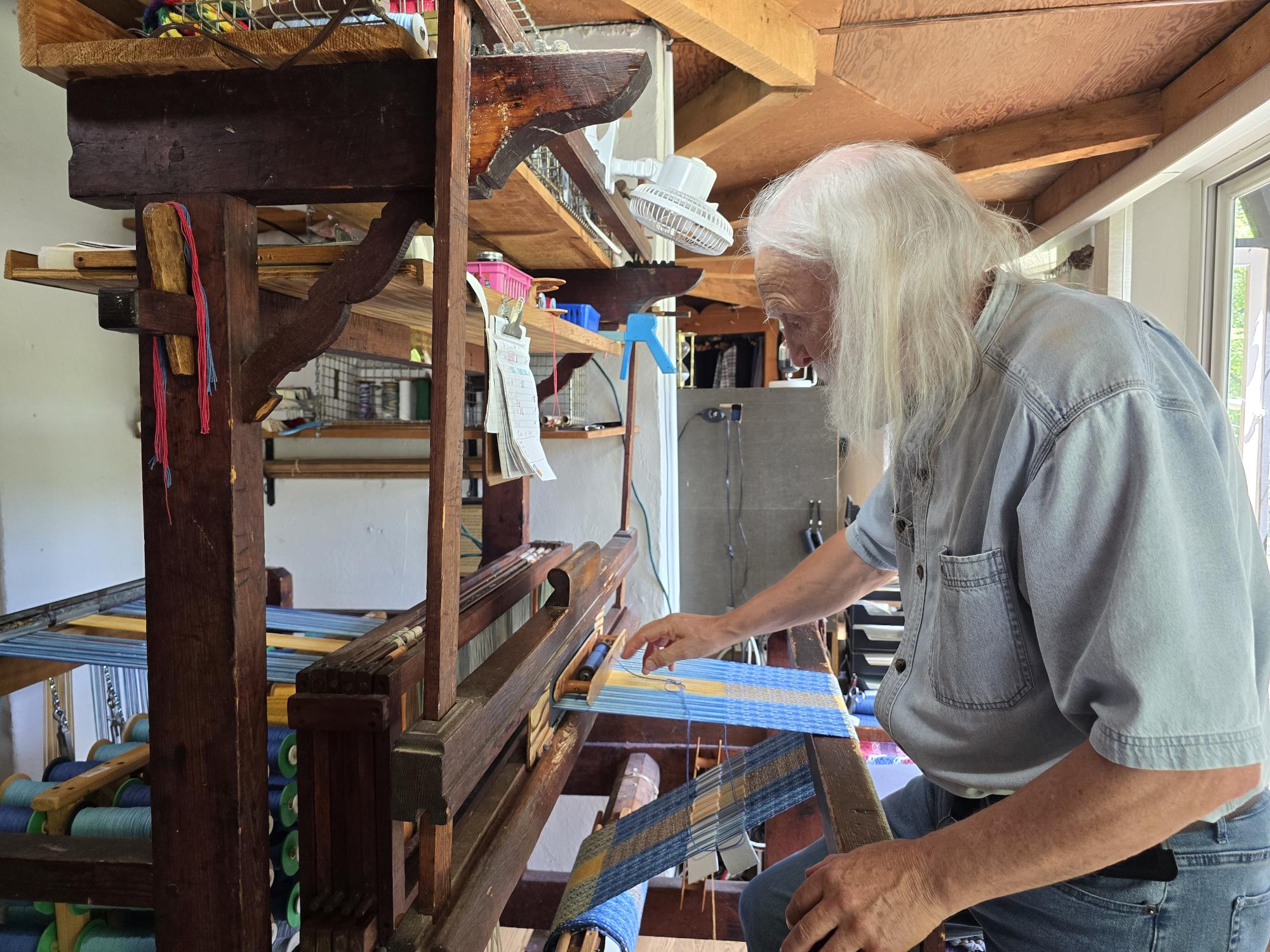 In a well-lit room, a person with long white hair skillfully weaves on an antique loom. Wearing a denim shirt, they concentrate deeply on their craft using 100% cotton threads, much like those used in the creation of the Flag: Flag on Hanger - Sweden.