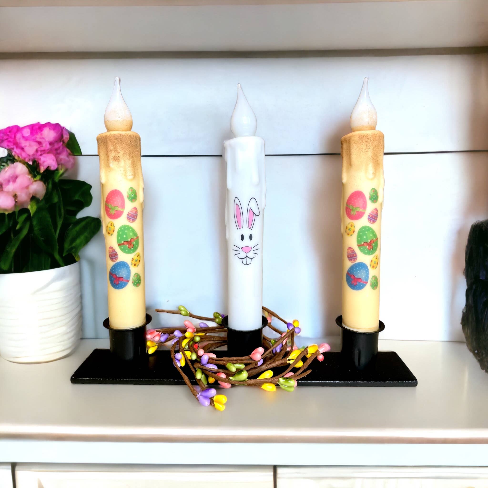 The Triple Wrought Iron Taper Holder elegantly displays three decorative candles with Easter designs, encircled by a floral string. This delightful centerpiece, accented by a pink potted plant on the left, adds festive sophistication to any shelf.