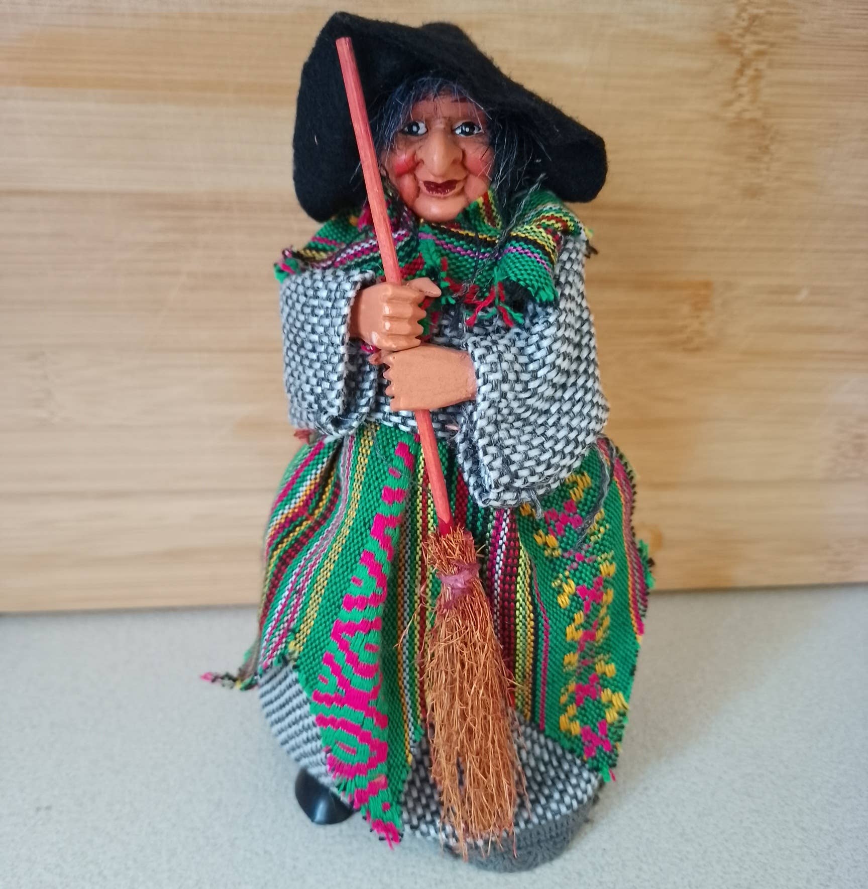 The Figurine: Yasmina Green Kitchen Witch 7" Tall features a handmade doll with a black hat, colorful shawl, and broom, displayed on a light surface with a wooden background.