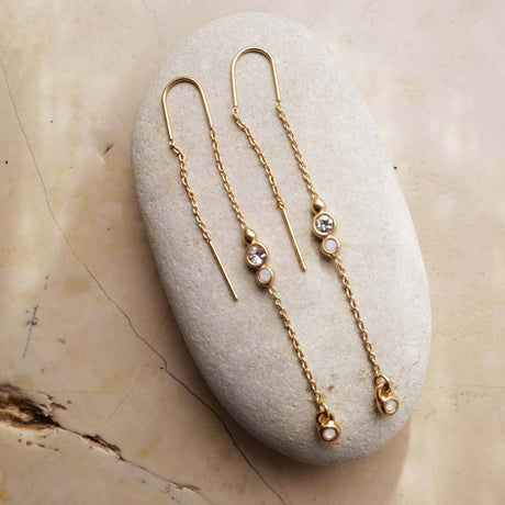 The A&C Oslo Long Crystal Chain Gold Earrings, featuring small round white stones and clear gems, are displayed on a smooth oval stone against a marble background, highlighting the elegance of organic jewelry design.