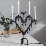 The Star Trading Metal Candlestick El-Tupp, a black heart-shaped candelabra with four lit candles, stands on a light table beside a glass vase with green foliage, creating a cozy Christmas atmosphere.