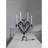 The Star Trading Metal Candlestick El-Tupp, featuring five lit white candles, stands on a light wooden table in a minimal gray room, bringing a subtle Christmas touch.